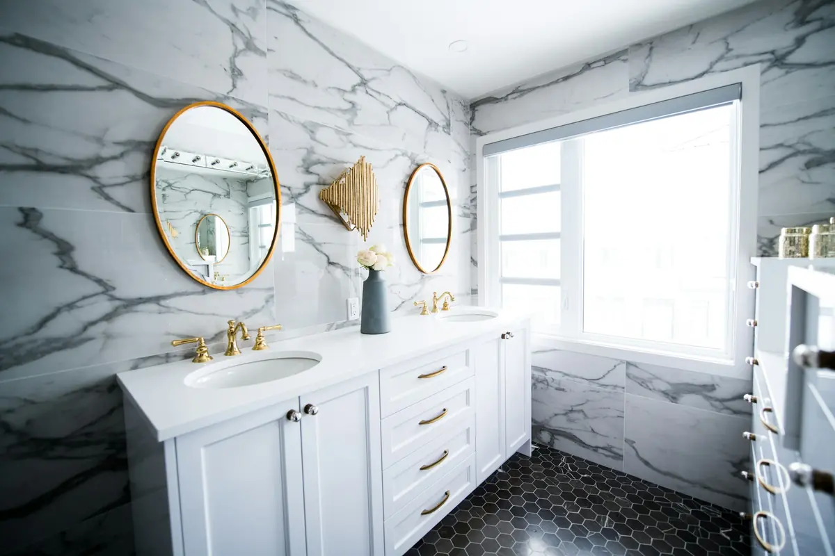 Marble-clad bathroom with white double vanity, brass faucets, round brass mirrors and hexagon floor tile