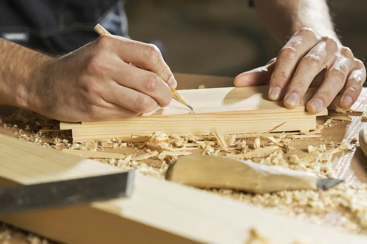 Carpenter's hands marking a wood board with a pencil beside a chisel amid fresh shavings