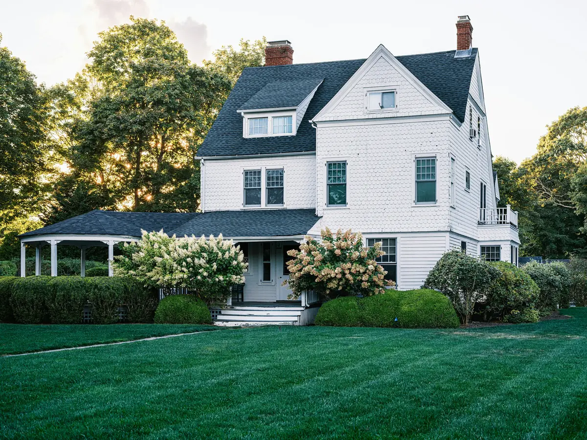 White shingle-style house with dark roof, wraparound porch, blooming hydrangeas and freshly striped lawn