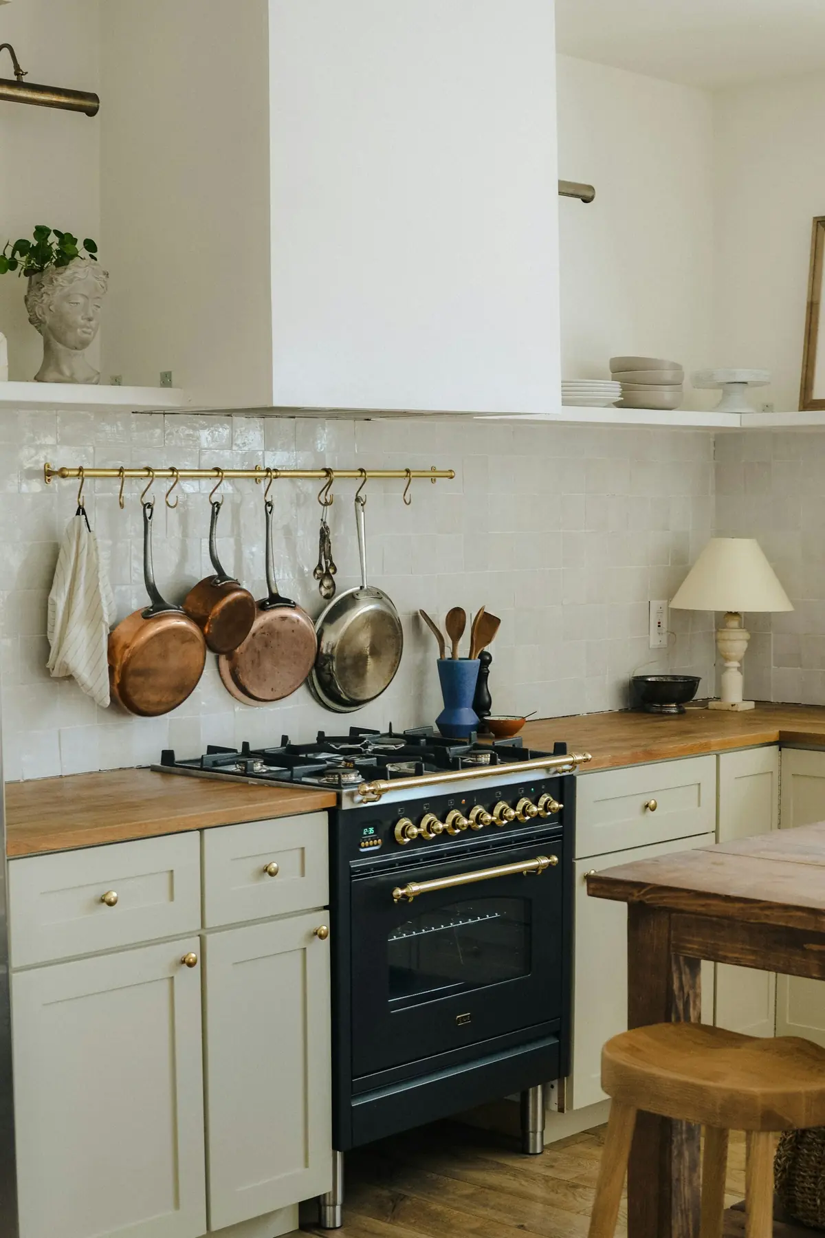 Kitchen corner with navy range, copper pots on a brass rail, white shaker cabinets, butcher-block counters